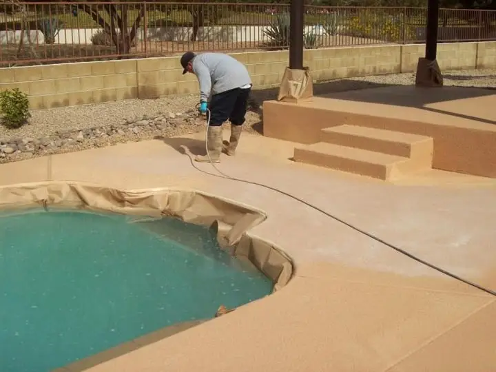 A crew member spraying coating onto a pool deck, the pool masked off with paper