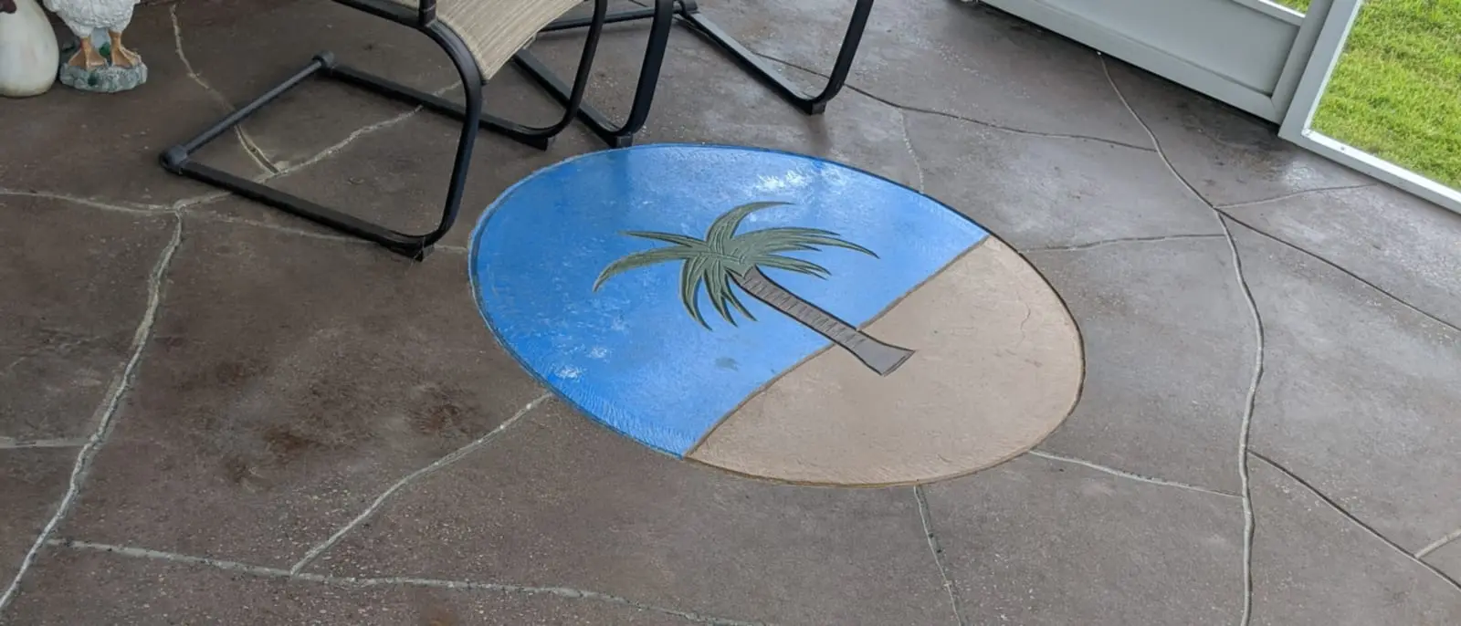 A stained and scored concrete patio with a blue and white palm-tree medallion set into the slab, chairs at its edge
