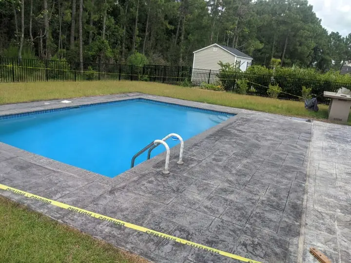 A blue swimming pool bordered by a wide grey stamped concrete deck and lawn