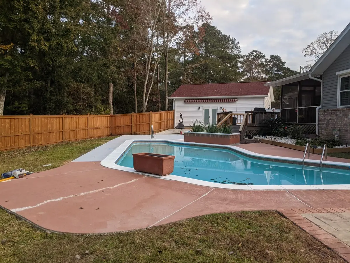 A backyard swimming pool surrounded by a broad reddish-brown stamped concrete deck