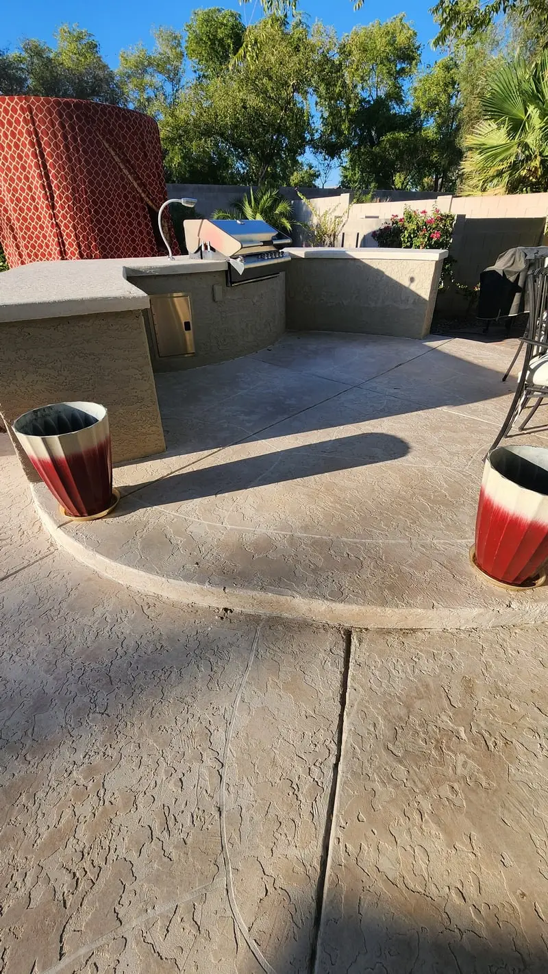 A curved stamped concrete patio wrapping an outdoor kitchen, palms and bougainvillea behind it under a blue Arizona sky