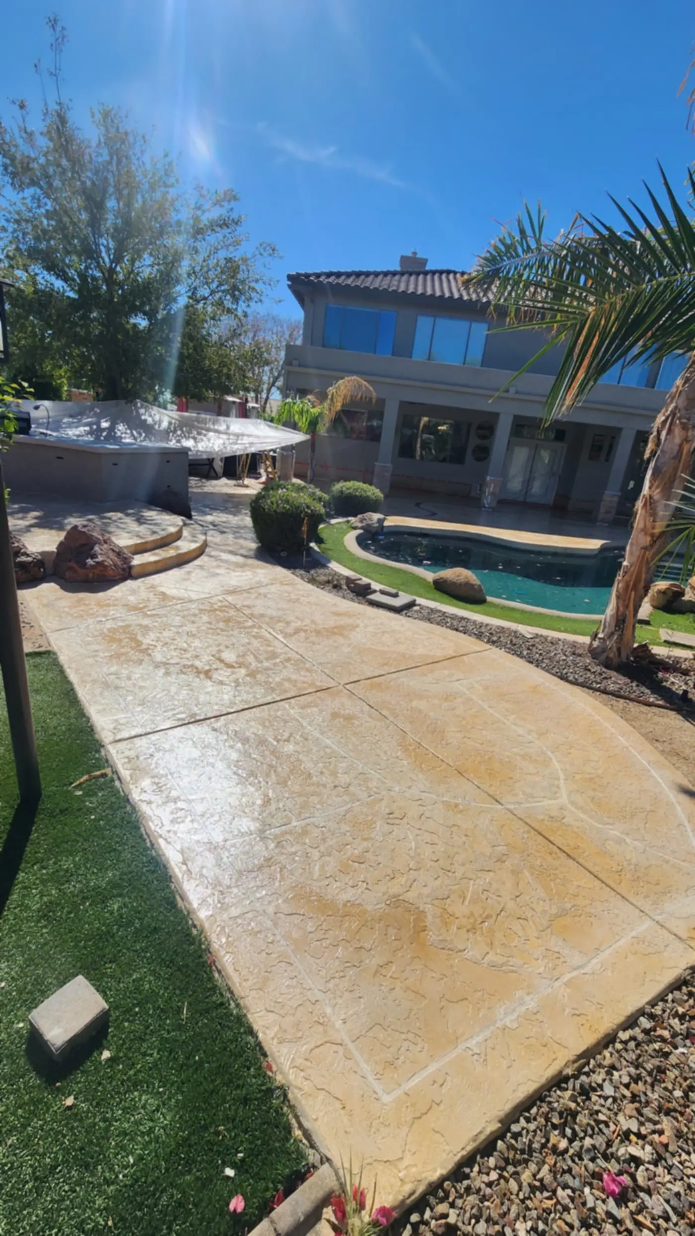 A wide gold-toned stamped concrete pool deck sweeping past a palm toward a pool and a two-storey stucco home under a clear desert sky